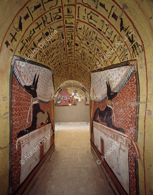 Pasillo de entrada de la tumba de Pashedu. A ambos lados se representa al dios Anubis en forma de chacal, descansando sobre un pedestal. Las dos imágenes son reflejos especulares la una de la otra. Entre sus patas traseras, Anubis sostiene un azote.